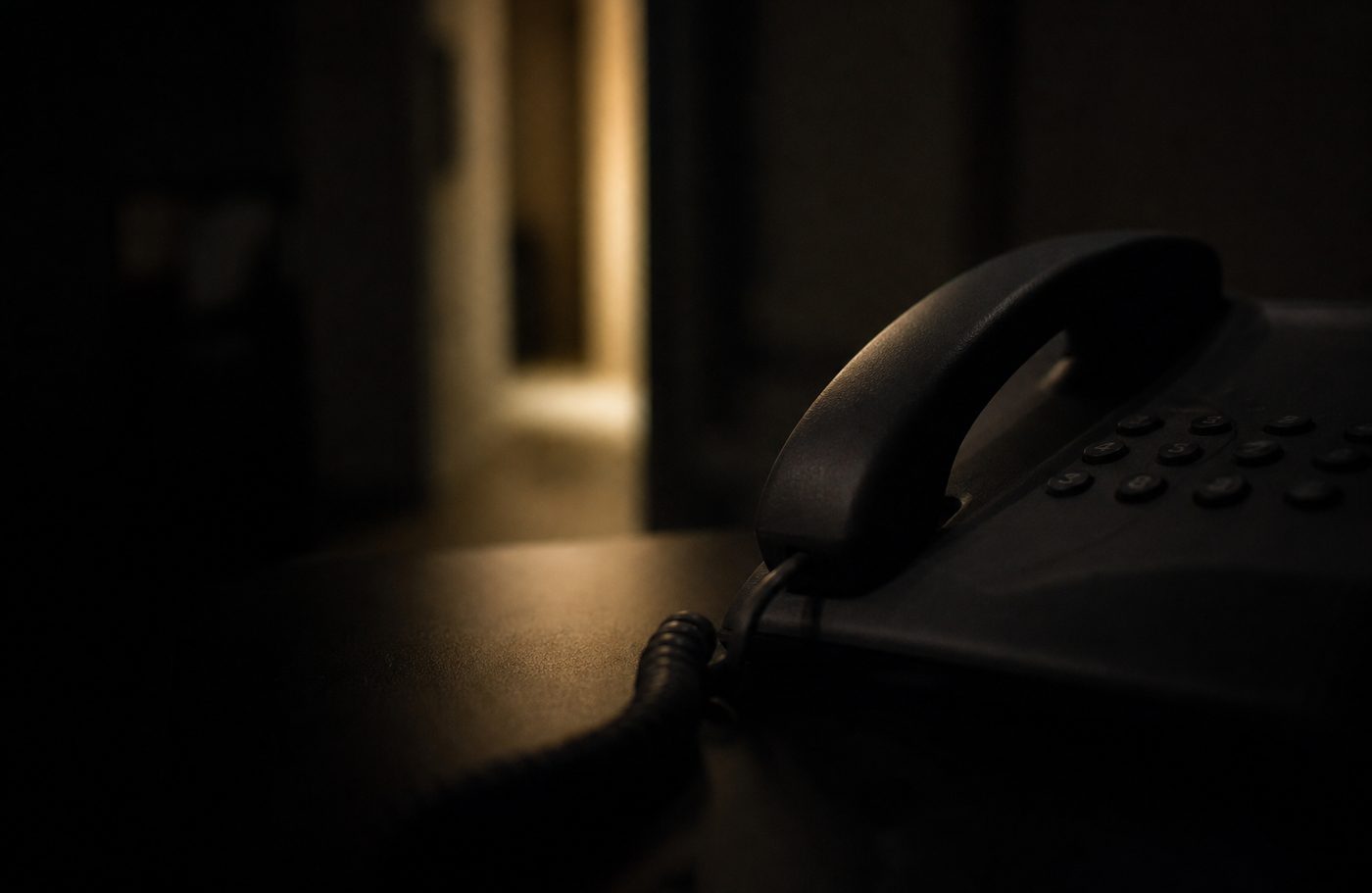 Desk phone handset lit by a doorway in a dark office, representing after-hours inbound restoration and HVAC calls